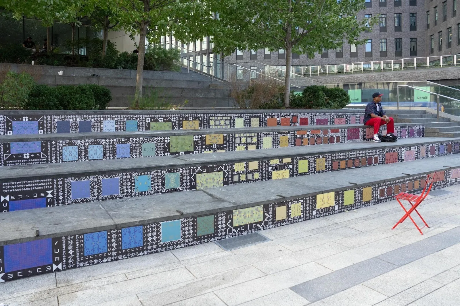 People sitting on black organically shaped benches with dreamscape markings in a public plaza. Some people are sitting int he background eating and talking to each other, The slopes and peaks of the benches are algorithmically generated from dreams.