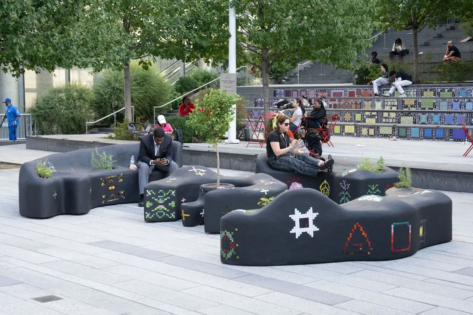 People sitting on black organically shaped benches with dreamscape markings in a public plaza. Some people are sitting int he background eating and talking to each other, The slopes and peaks of the benches are algorithmically generated from dreams.