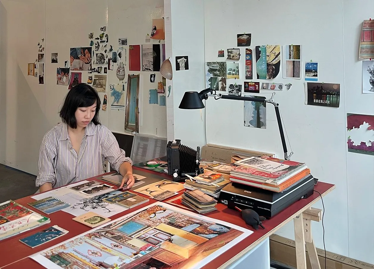 Chinese female artist with short hair wearing striped collar button down shirt in front of a desk full of photographic references and Architectural archival images, photographs on the wall, silver art projects Studio space Xi Li photography.