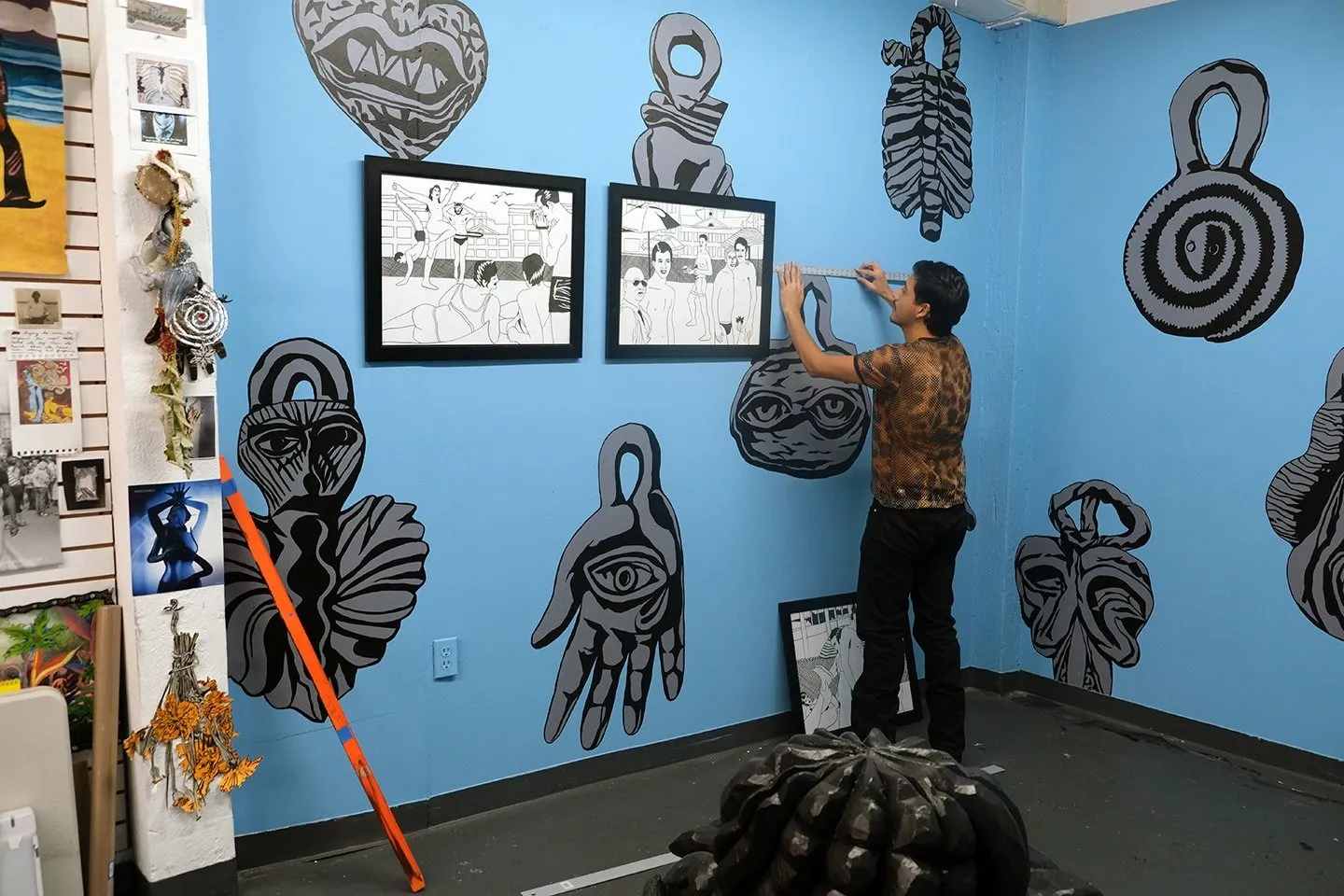 Mexican American artist Miguel Martinez putting a ruler against blue steel wall with traditional Mexican imagery, Studio portrait with sketches pinned to the wall and drawings layered on top mural.