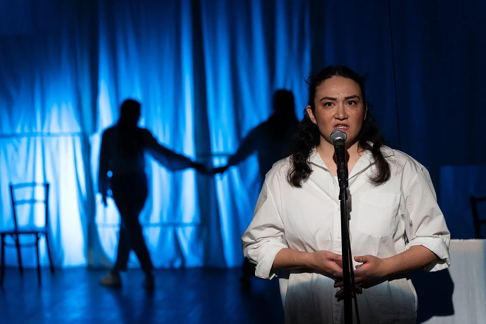 One woman in white in front of a microphone speaking on stage against blue curtain background with two Shadows, The Brick presents Lucky Breaks by Yevgenia Belorusets.