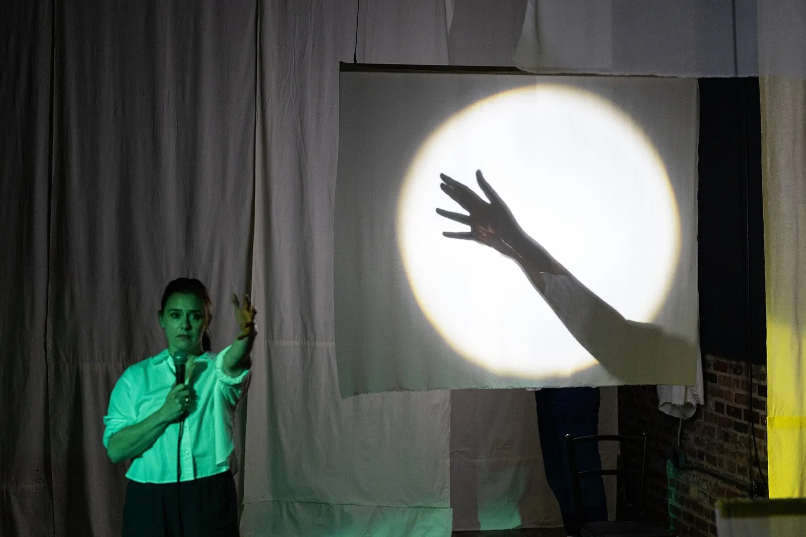 One woman holding microphone with hand reaching out, she wears white blouse and is lit by green light, projection screen behind her casts shadow of her hand amid bright white ring of light, The Brick presents Lucky Breaks by Yevgenia Belorusets.