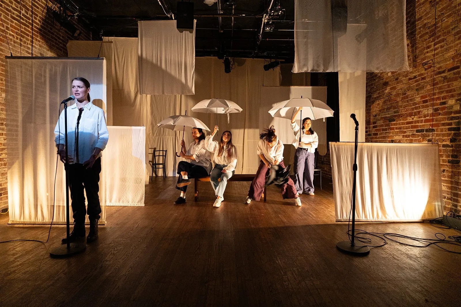 Five actresses wearing white shirt on the other stage holding white umbrella against the curtain backdrop, The Brick presents Lucky Breaks by Yevgenia Belorusets, one speaks into microphone while others sit on bench.