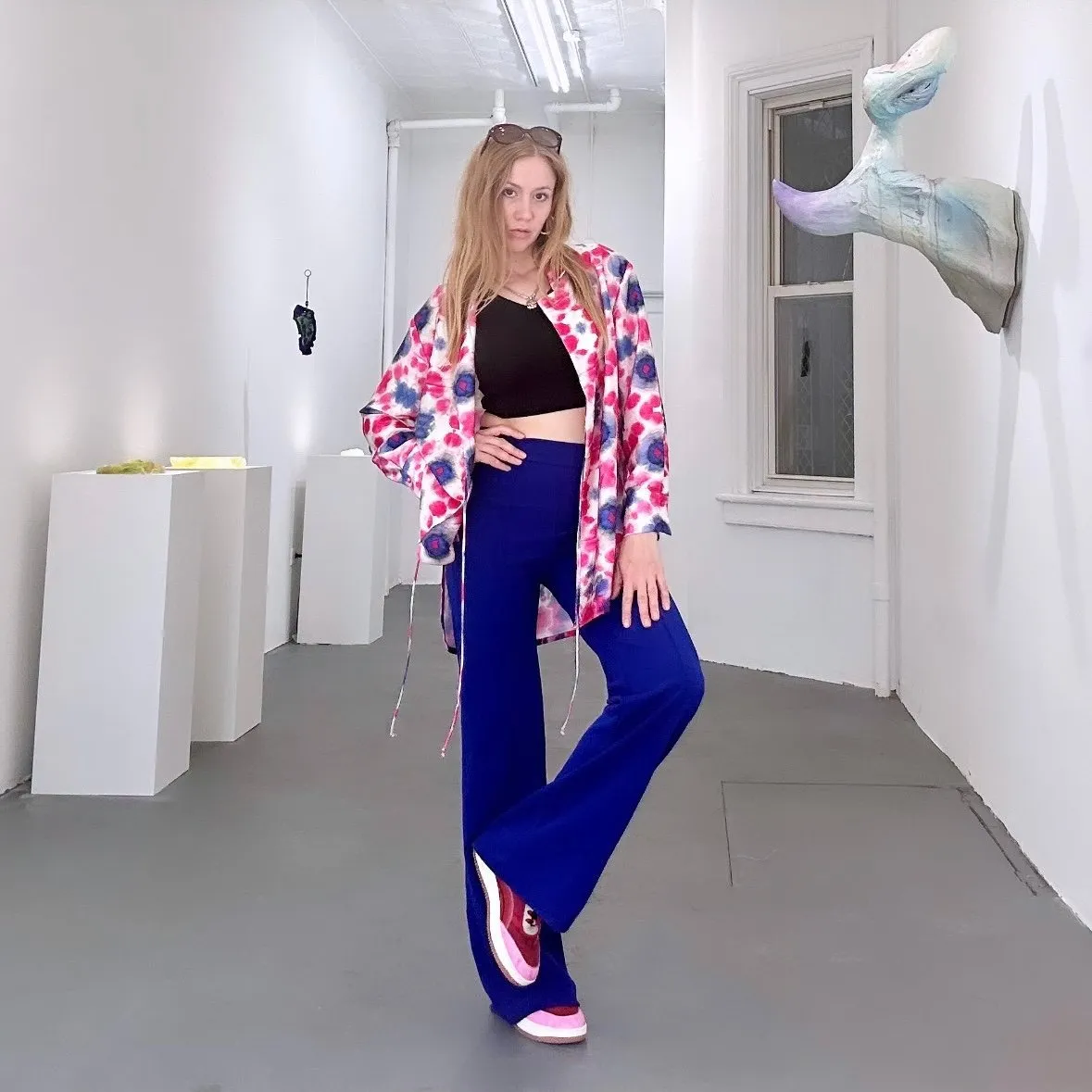 Blonde woman wearing pink and blue shirt, blue pants, and sneakers in front of her work, Portrait of Lydia Nobles at her solo exhibition, Body Lies, at KAPOW in 2024, work of Lydia Nobles reproductive rights and sexuality sculpture.