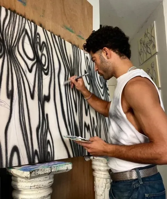 Rubeen Salem paints in his art studio on the Lower East Side, male palestinian artist wearing white tank top and denim jeans painting black wave lines on white canvas.