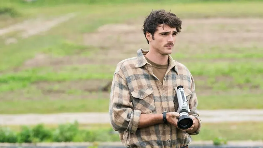 Jacob elordi in checkered brown shirt holding camera looking serious in grassland, still from oh canada.