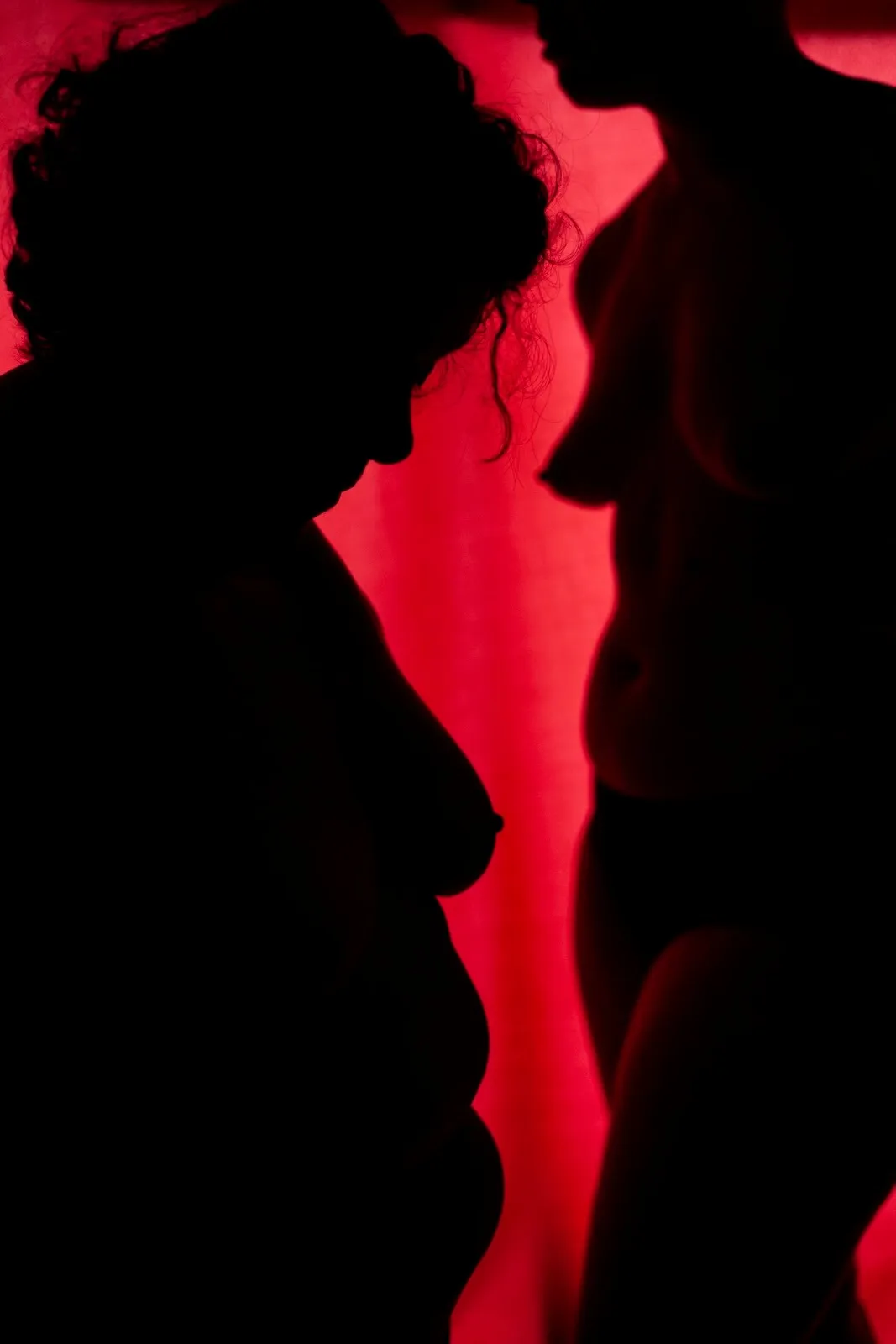 The dark silhouettes of two naked women standing against luminous red background, from Jeanette Spicer's intimate photography series, to the end of the earth.