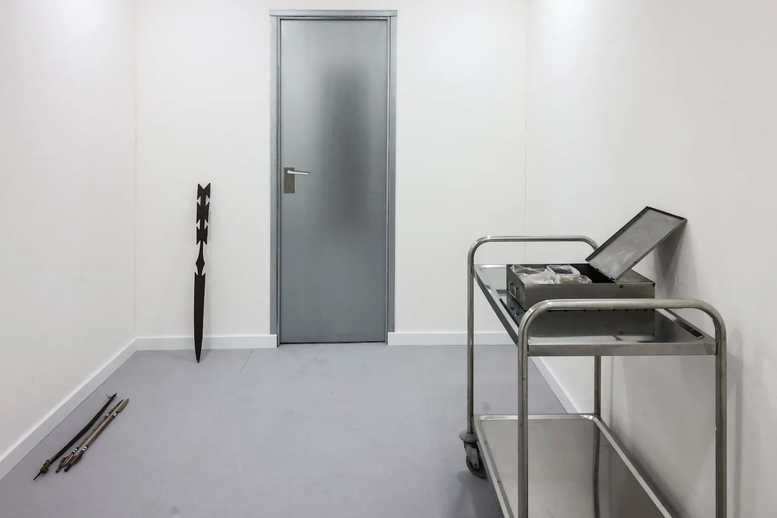 Room with white walls and steel door, spear-like sacred brazilian indigenous artifacts leaning against the wall and on the floor, a shelf with an open container on the right, Lucas Memmola, Paradise Bank, antechamber, art installation, Rome.