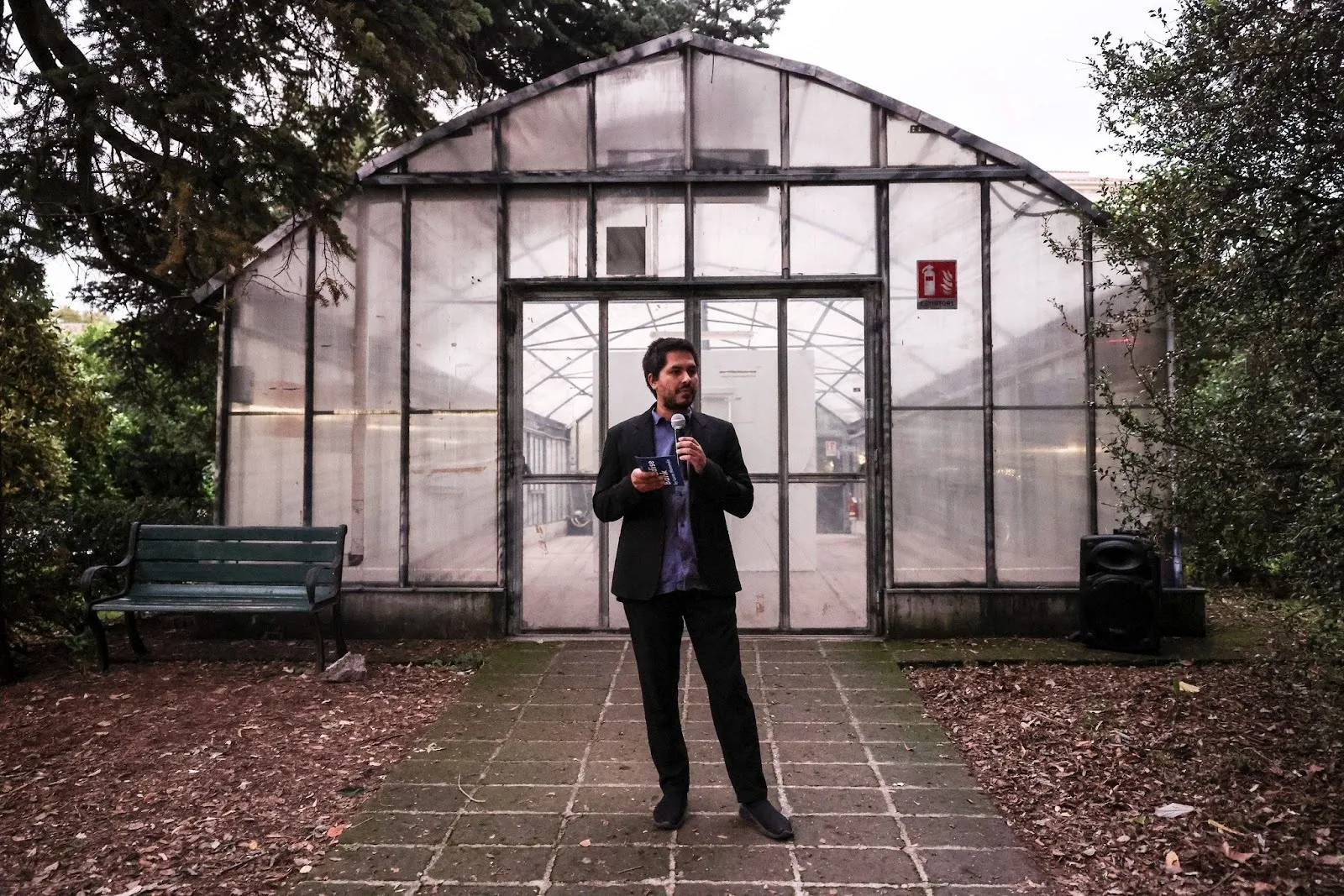 Man in suit standing in front of glass greenhouse door in garden delivering speech, artist Lucas Memmola presenting paradise bank at the botanical garden in rome.