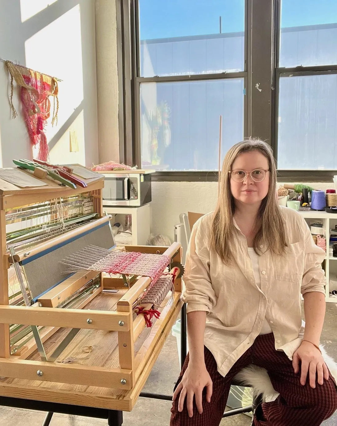 Ukrainian fiber textile artist Kathie Halfin sitting next to her loom with pink threads work in progress in artist studio in front of window.