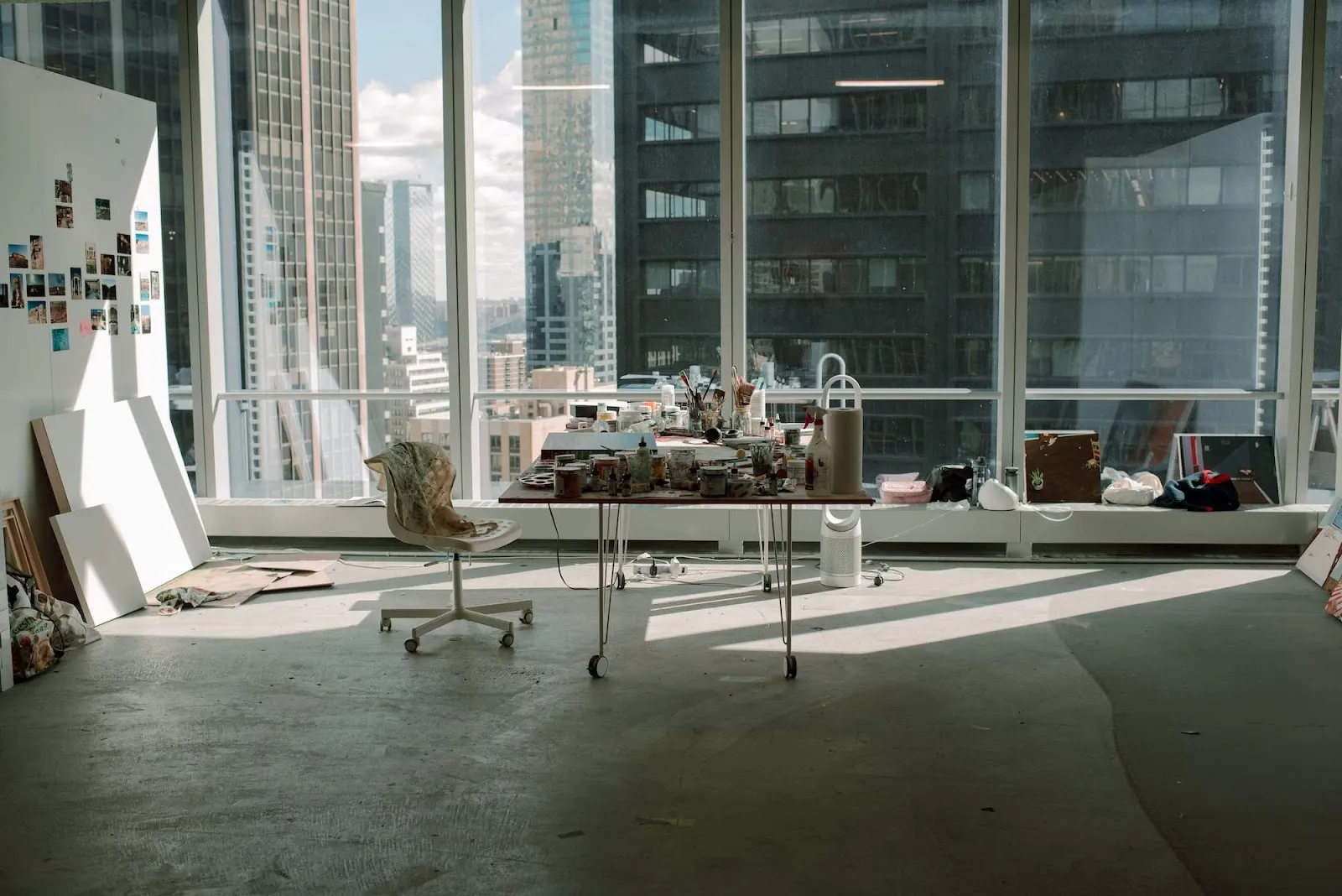 World trade center artist studio at silver art projects residency, table with paint and supplies in the center, huge floor-to-ceiling window overlooking skyscrapers, empty canvases and inspo photos, azadeh nia studio, iranian artist.