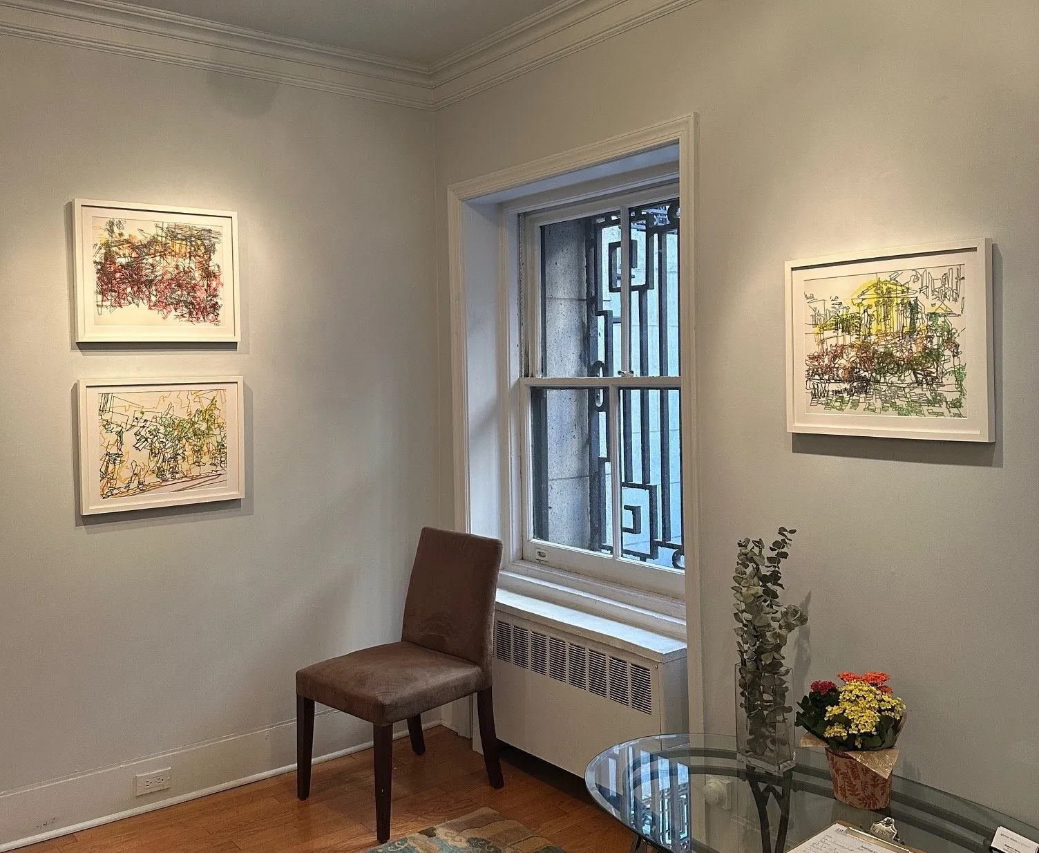 Three blind contour technique drawings of web-like lines rendering protest images, installed in office with chair, a clear table, and flowers, DFN projects out of line marjorie van cura solo exhibition.