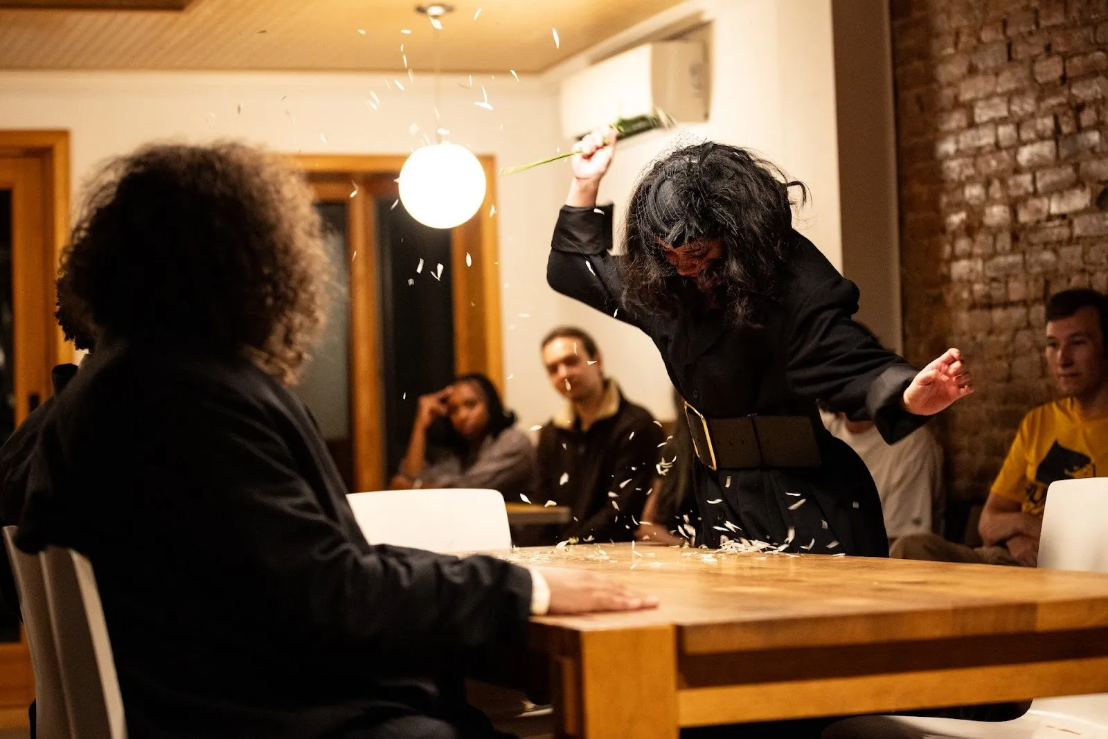 Actors on stage around deskm luis feliciano faces izabel mar, a woman in all black stabbing a flower with loose petals, family off-broadway theater by celine song directed by alec duffy in hoi polloi and amanda james.
