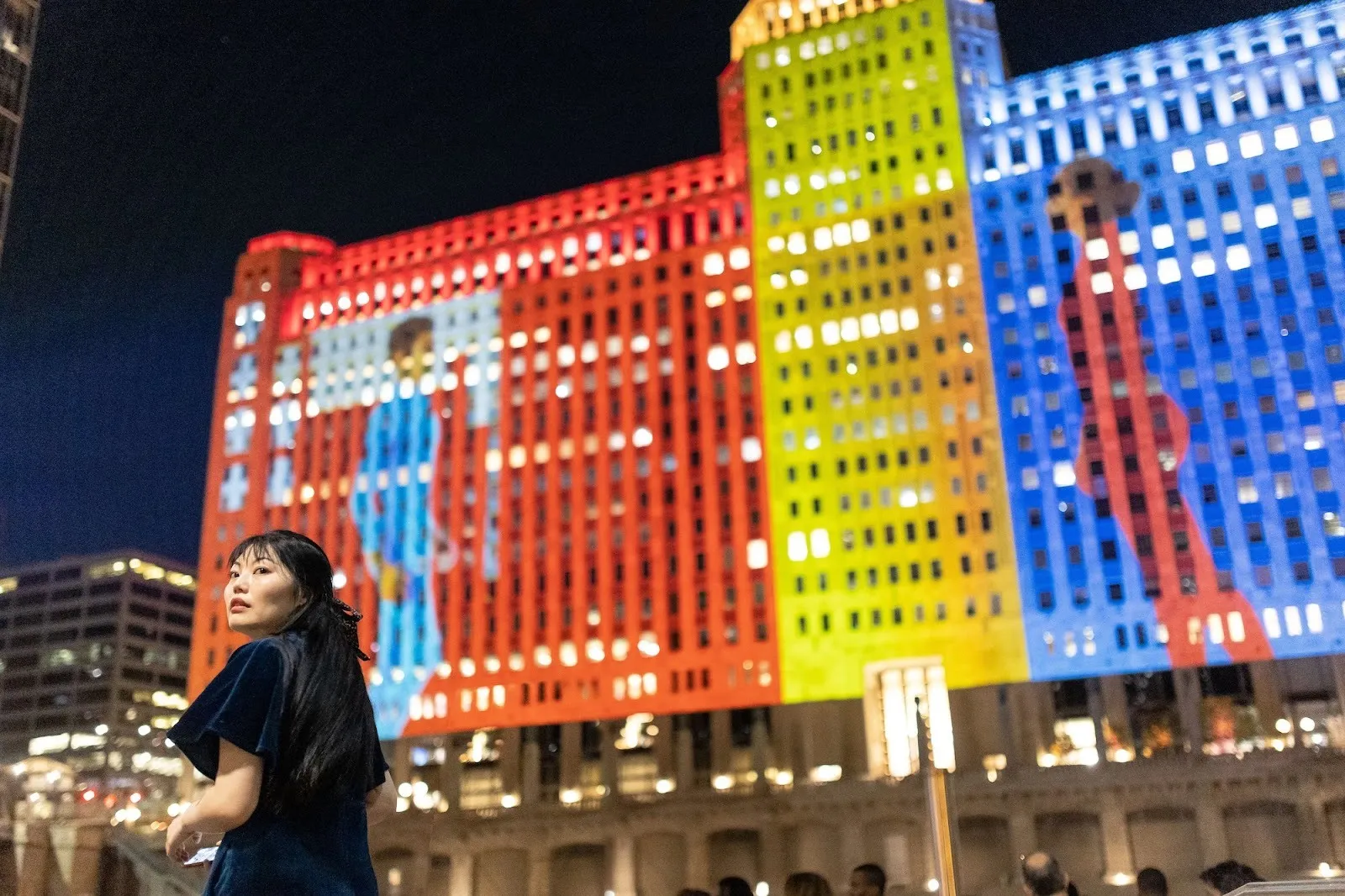 Chinese artist Yuge Zhou in front of red yellow blue video projection love letters at art of the mart in chicago, two dancers digital projection communicating with each other on building.