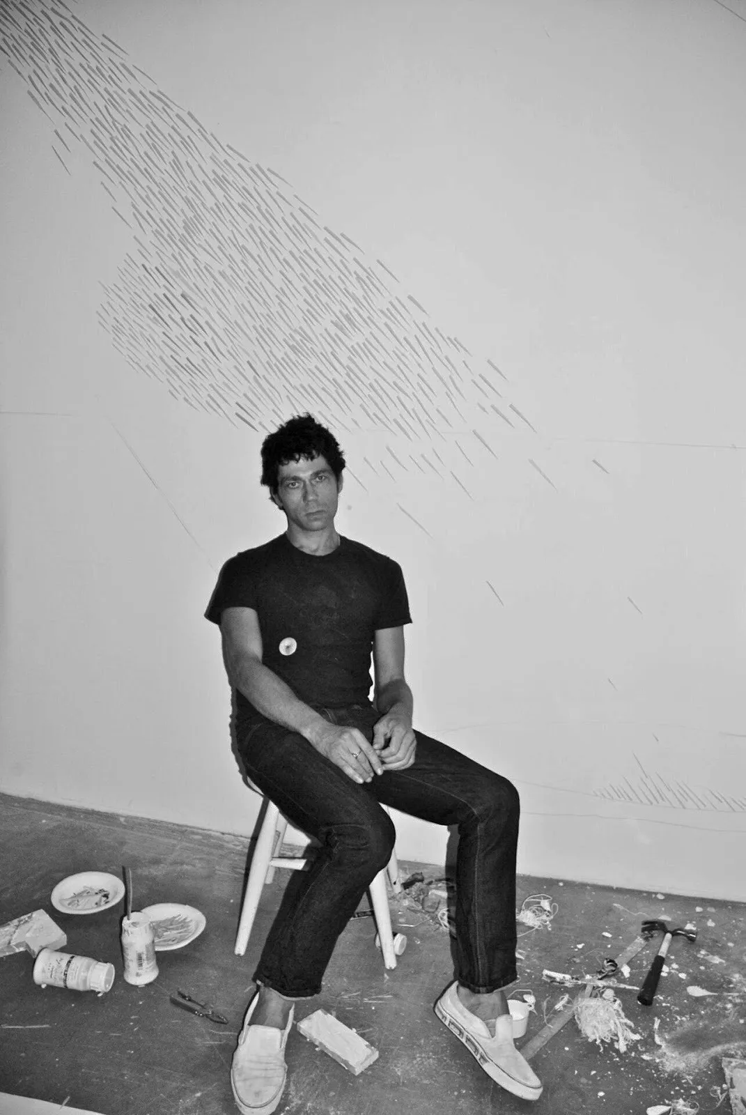 Man wearing black t-shirt and jeans sitting on artist stool in studio with paint around the floor, palo gallery happy land, british artist solo exhibition.