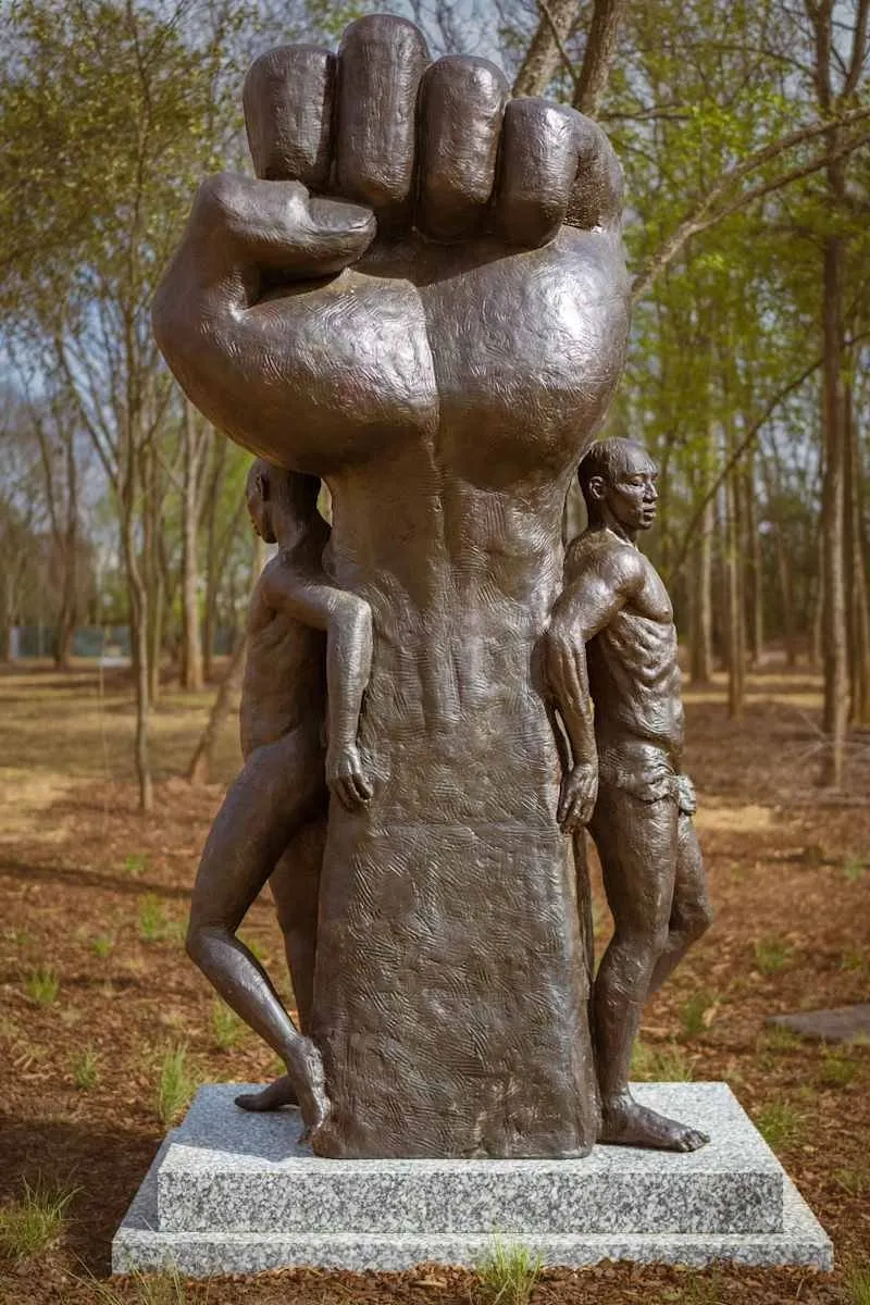 Bronze sculpture of two men standing next to larger than life fist, installed in a park, austen brantley power in the struggle, freedom monument sculpture park.
