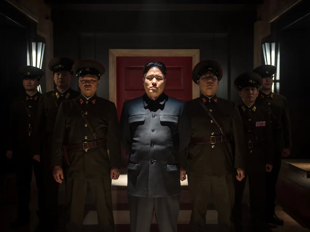 Actor Randall park playing north korean leader kim jong un in the 2014 film the interview, surrounded by security guards with eerie focused lighting, movie by seth rogan and james franco, review by lisa kwon.
