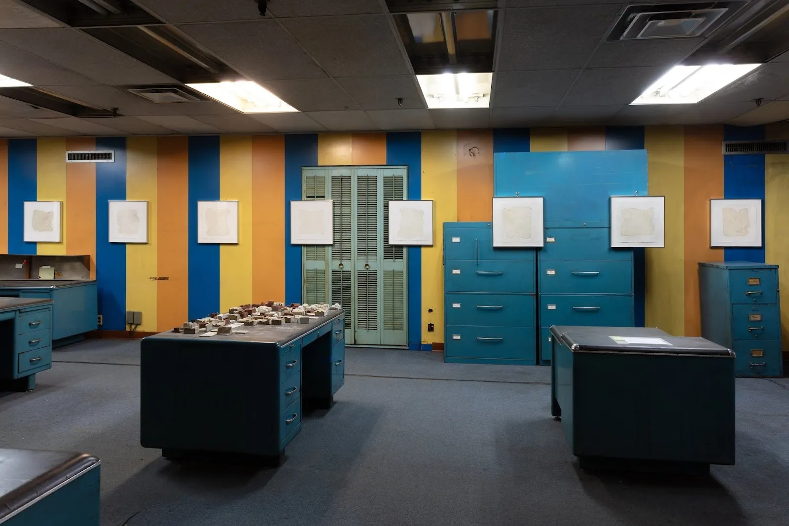 Installation view of an image of your labor hovers over me, group Exhibition at new uncanny Gallery curated by qingyuan deng, wall with yellow orange and blue stripes, blue metal cabinet, dominic palarchio, adriana furlong, christian amaya garcia.