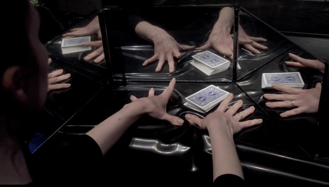 Two hands dealing cards on table reflected in three mirrors, still from taken by artificial surprise by jeanette andrews, photograph by derrick belcham.