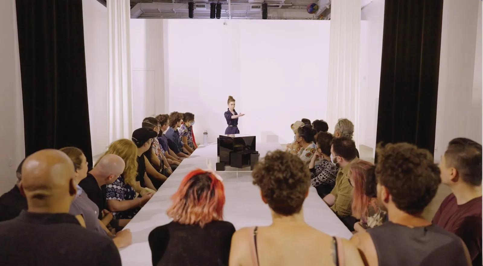 Magician Jeanette Andrews extending a hand in front of audience sitting around a white table at a performance, still from documentation of taken by artificial surprise, photograph by derrick belcham.