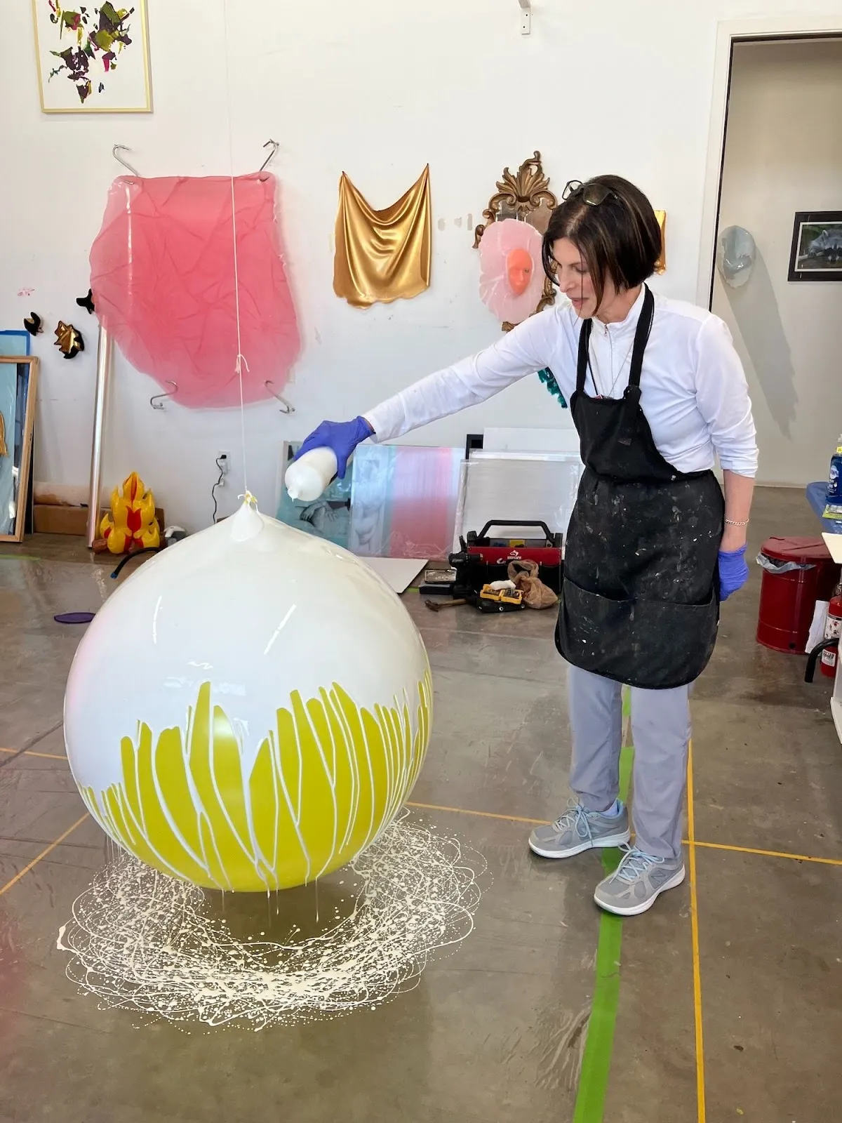 Sculpture artist Lesley Bodzy pouring white paint on huge yellow balloon stringed to the ceiling of artist studio, symbolic of beauty standards and femininity, interview with journalist xuezhu jenny wang.