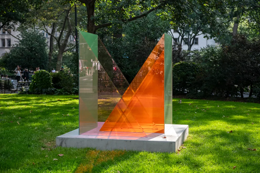 Intersecting panels of orange and green tinted glass form a sharp geometric sculpture on a sunlit lawn.