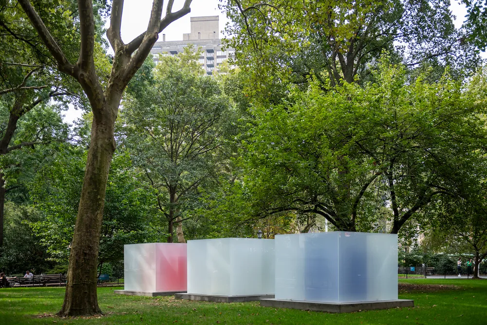 Three frosted glass cubes with subtle color gradients stand in a row beneath tall trees in a lush park.