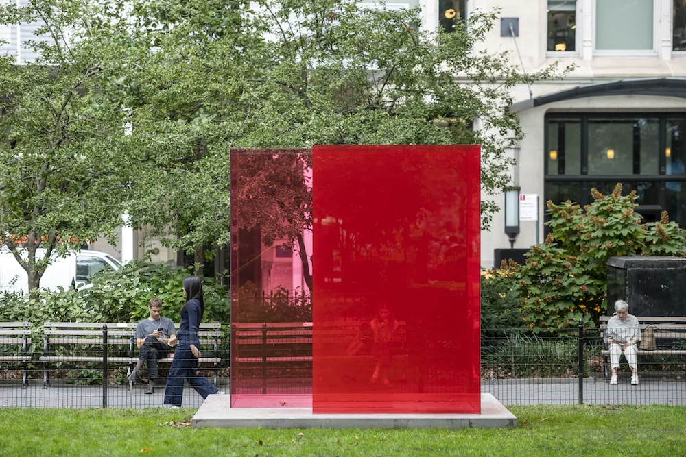 A bright red glass cube reflects pedestrians and park greenery, positioned near benches and surrounding foliage.