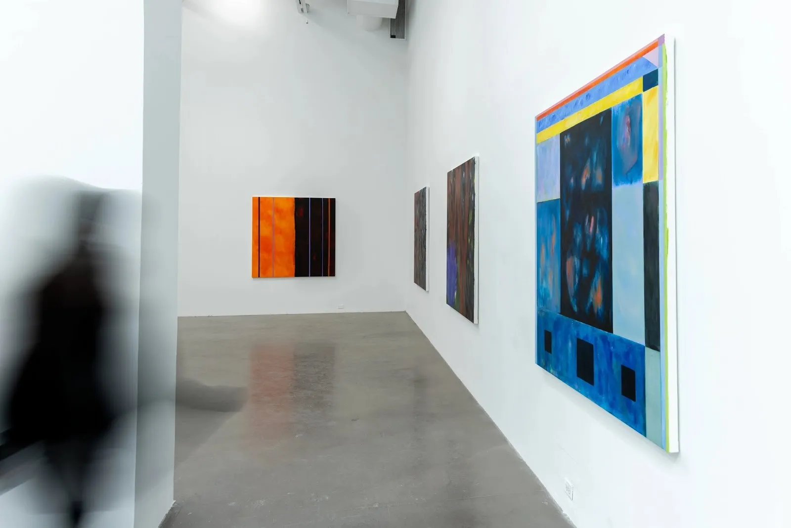 Gallery view with several abstract paintings in bold colors; a blurred figure moves past a blue painting in the foreground.