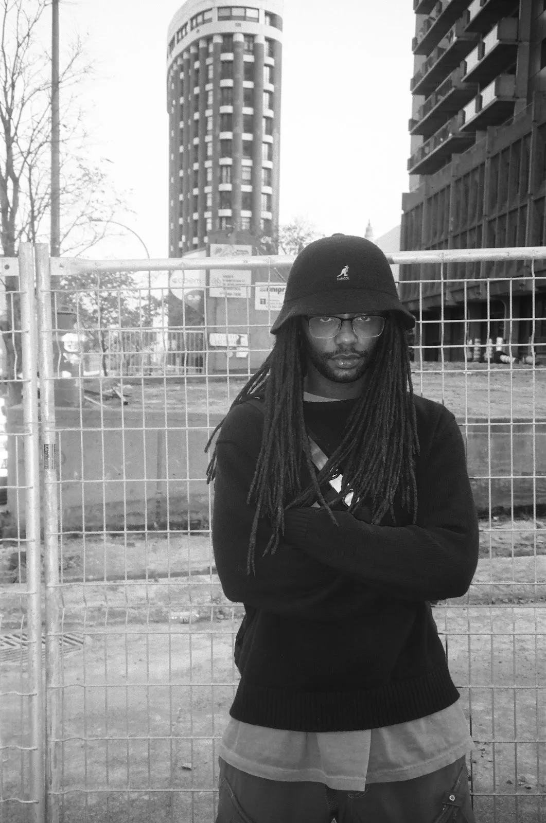 Musician DJ Gum.mp3 stands in front of fence and a park wearing bucket hat and black sweater, cinematic black and white portrait.