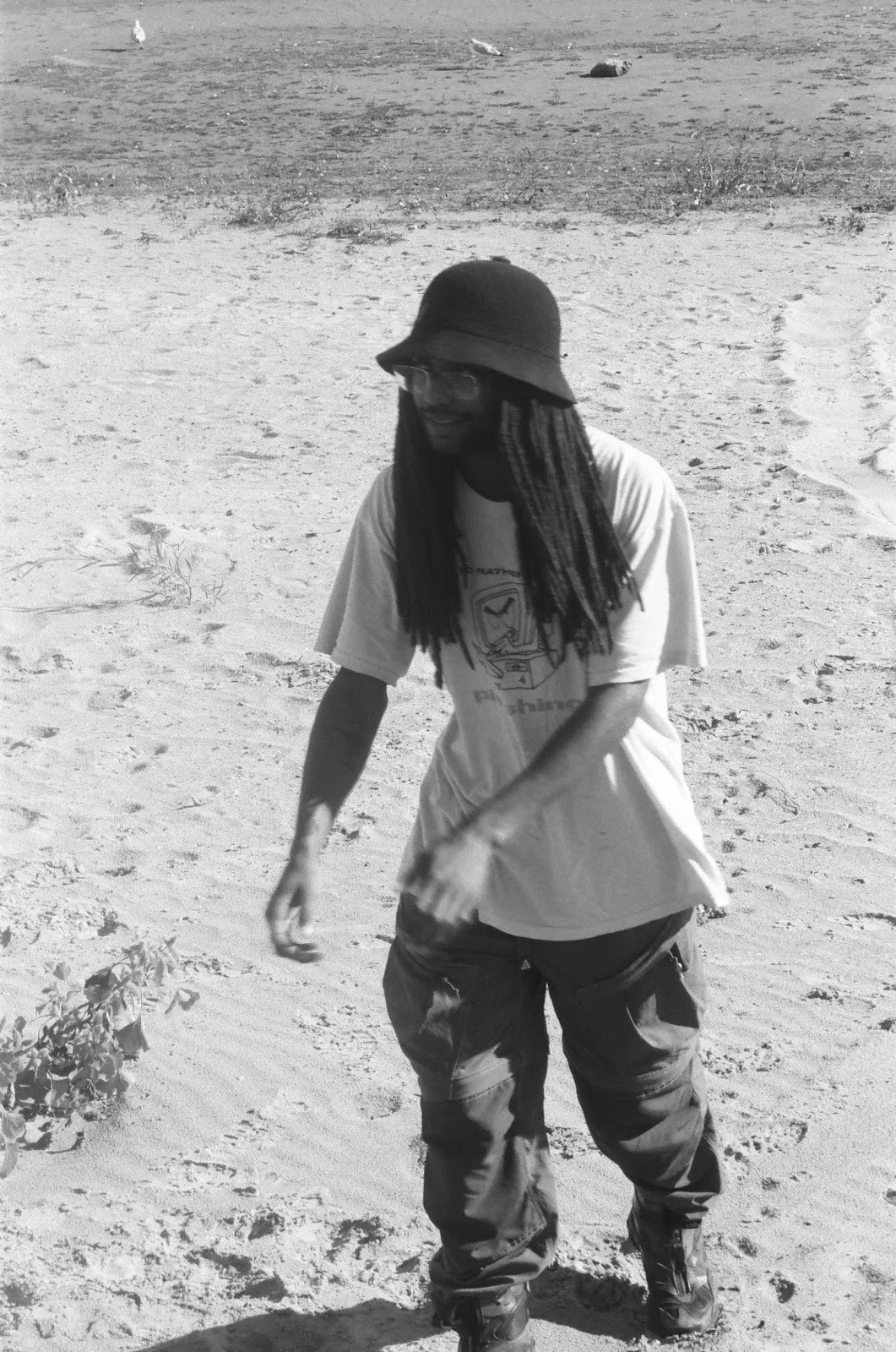 Musician DJ Gum.mp3 walking on sandy beach wearing white T-shirt and bucket hat swinging arms around, cinematic black and white portrait.