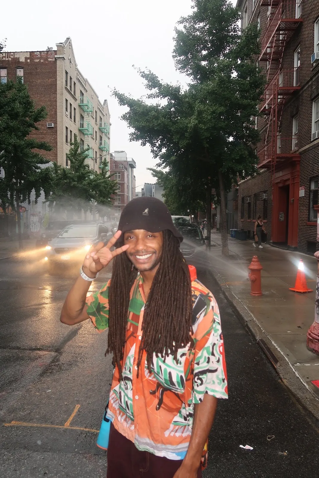 Musician DJ Gum.mp3 stands in the middle of New York Road wearing tie dye Tshirt and bucket hat with sprinkler behind him, holding hand in victory sign.