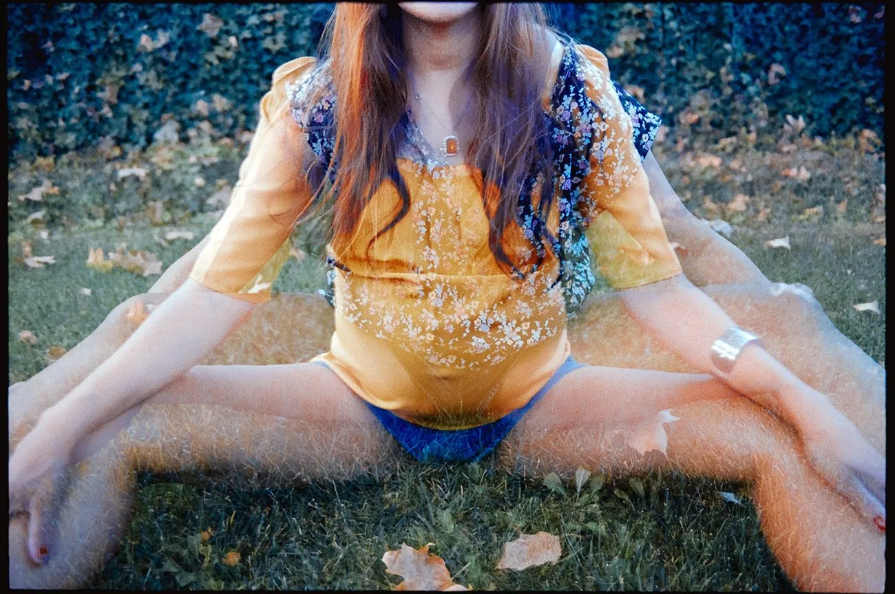 Color analog photo of girl in orange top and blue underwear sitting on grass with pregnant belly, photographers Amber Maalouf and Ashlie Chavez.