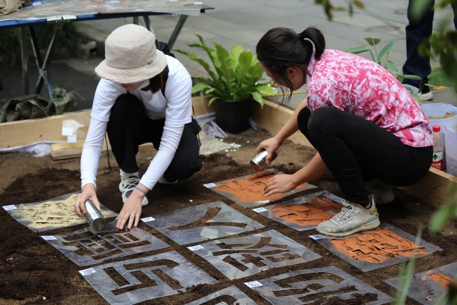 Two women squatting side by side on soil-covered garden spraying paint over Chinese character templates, performance art.