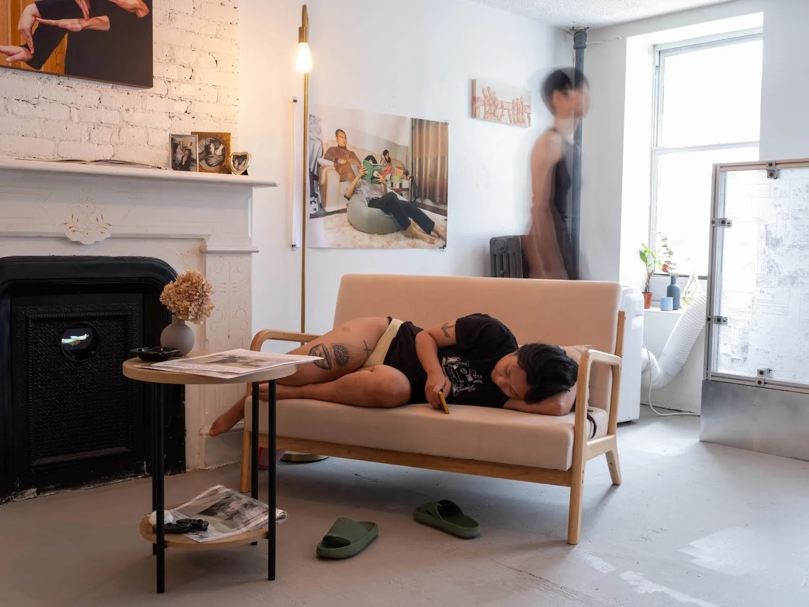 Asian girl curled up in couch scrolling on phone, shadow of man rapidly walking behind her in living room with photographs all over and a lit lamp, bandaged moments at Tutu Gallery.