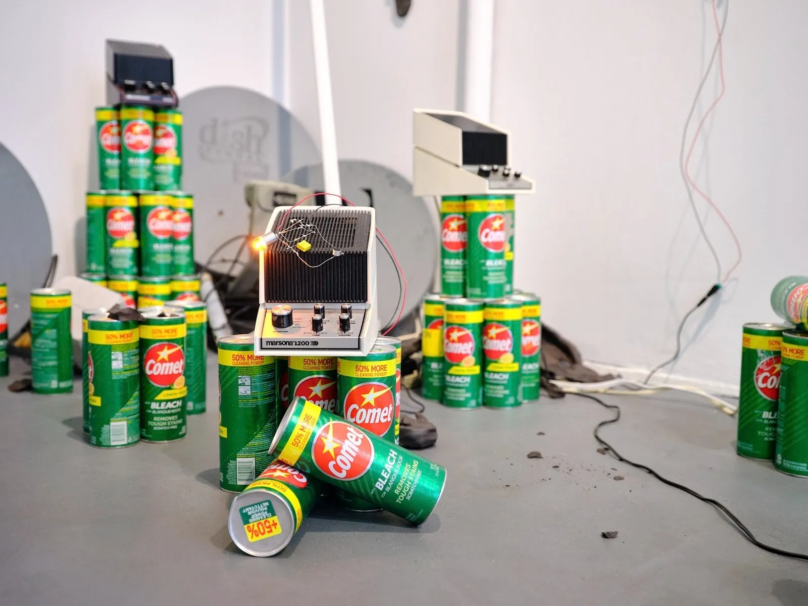 Green bleach cannisters stacked on floor with Marsona sleep aids noise machine, circuits, Black ears and sattelite dishes in white cube gallery, Char Jere, peripheral belonging group show at GHOSTMACHINE.