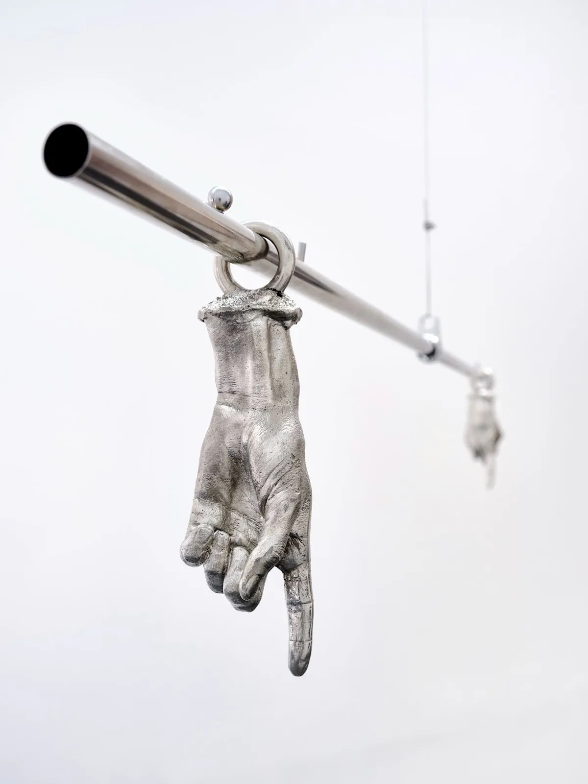 Two pewter hands with index finger pointing down dangling from metal bar, Guillermo Rodriguez, double figure at peripheral belonging.