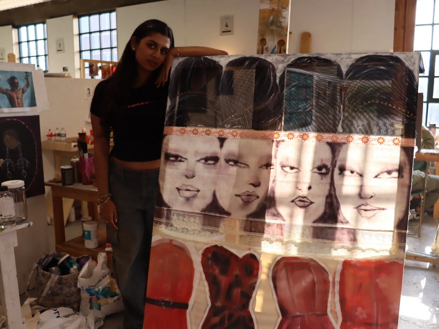 Indian artist Maithili Chaturvedi in her studio. Long-haired woman with black T-shirt and denim skirt next to translucent painting with four women faces and red dresses.