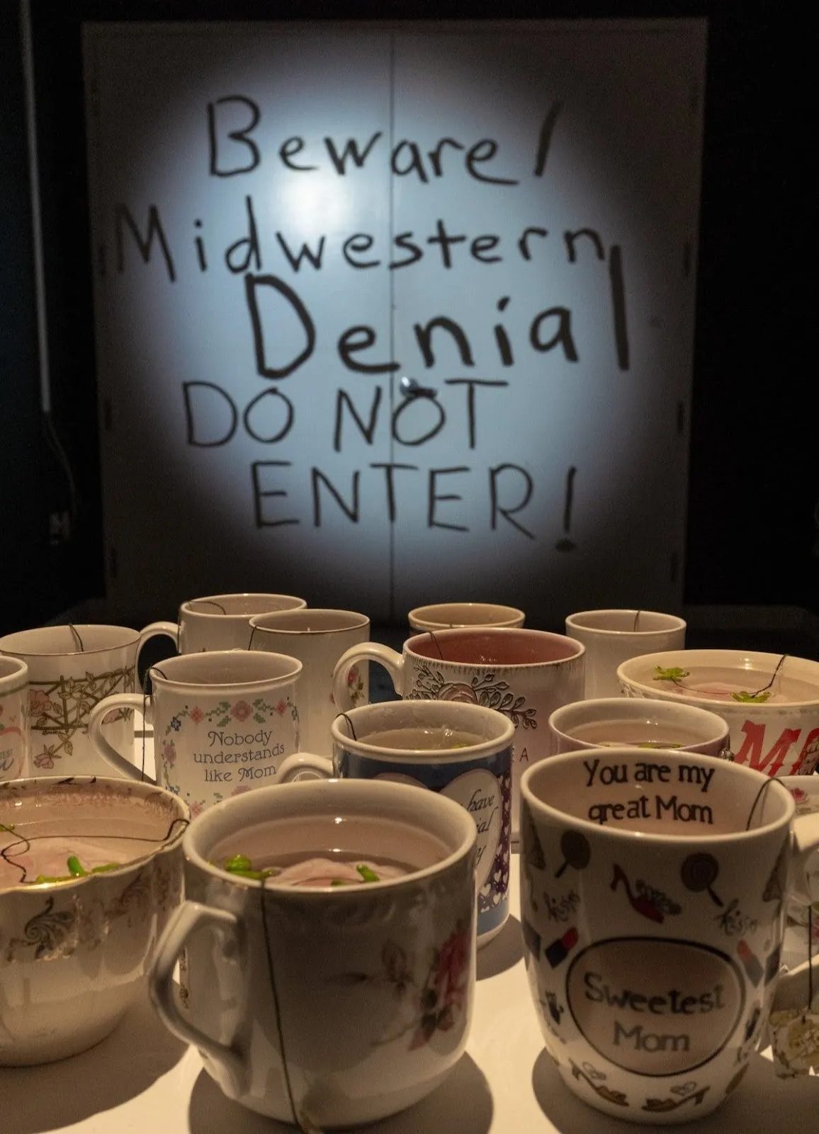 Many teacups in front of wall projection that says beware Midwestern denial do not enter, immersive exhibition gallery Philadelphia, the fabric workshop and Museum the Rose Garden John Jarboe.