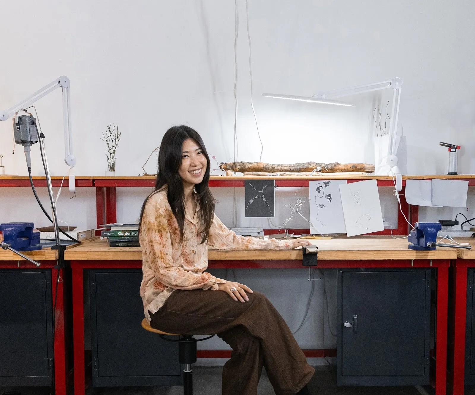 Smiling Asian woman designer with long hair sitting next to jewelry desk, wearing orange floral print blouse and brown pants. Aeri Go.