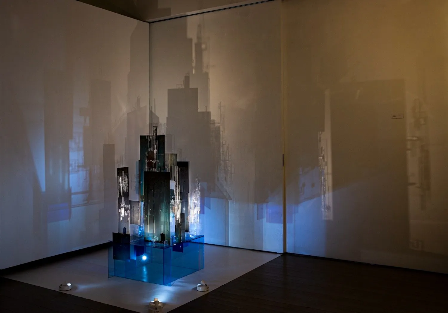 Black and blue acrylic sheets rectangle printed with buildings and cityscape photographs, stacked into tower-like sculpture in dark room with lights Illuminating from underneath to project shadows on the walls.