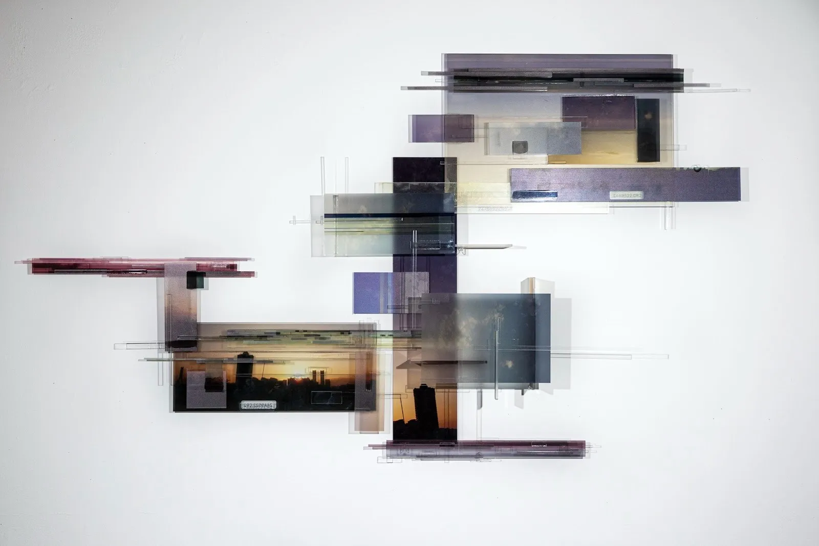 Layers of purple black and orange acrylic rectangle sheets layered in sculptor form, printed with city landscape and sunset against white wall background, Jingyao Huang. Chaotic Data Fences, Baraka, and the Cityscape #1.