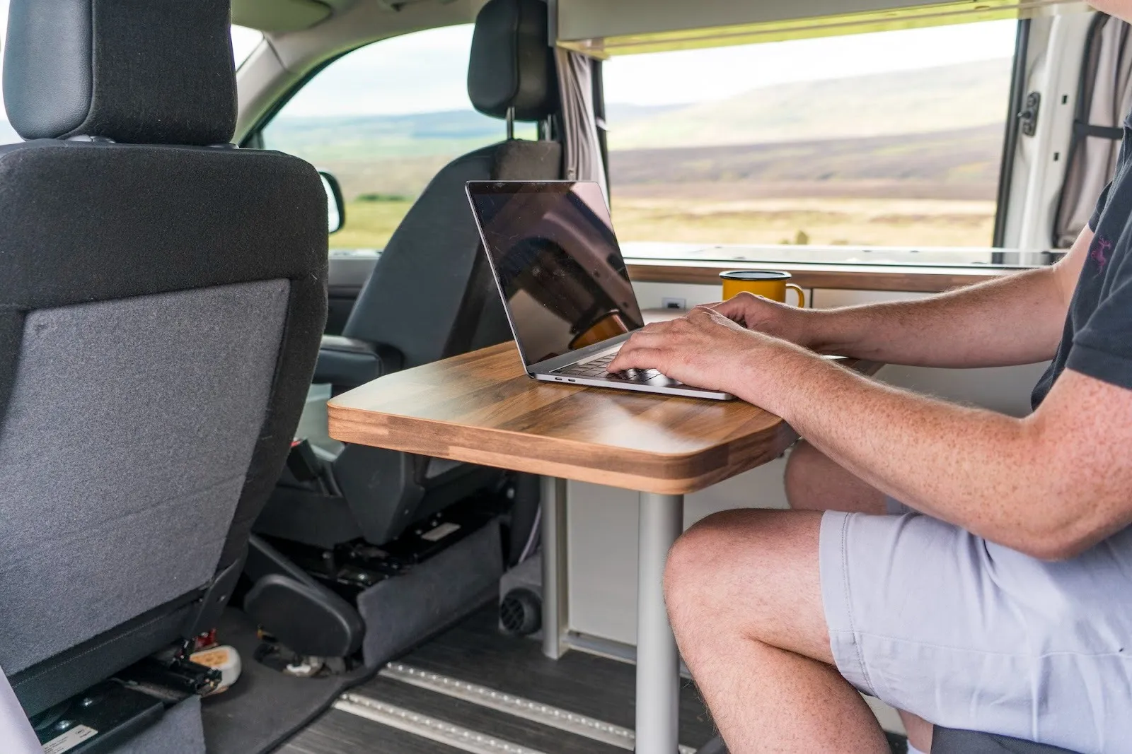 Photograph of a man working in the back of a campervan in the middle of the countryside. He is working on his laptop and has a cup of tea beside him. Hills and fields can be seen out of the campervan window, digital nomad visa atreya mathur.