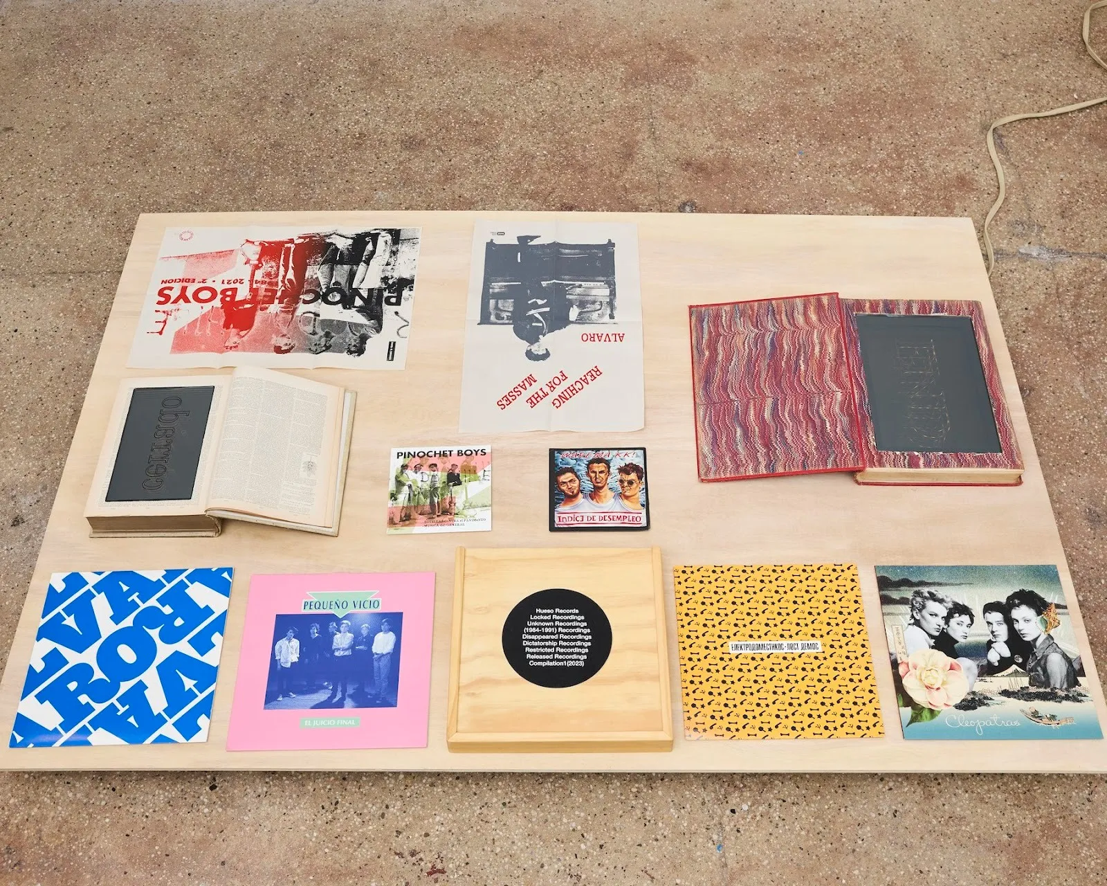 Wood board with pictures, books, notebooks, and postcards on gallery carpet, installation view of Iván Navarro solo exhibition Small Talk at Miriam Gallery, chilean political art reviewed by emma jones, impulse magazine.