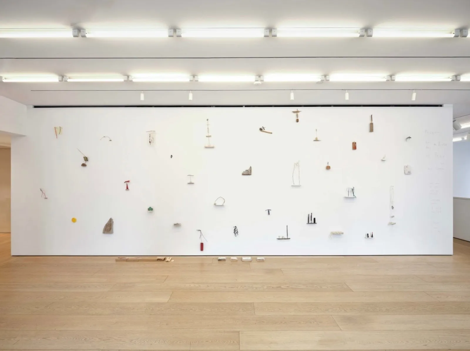 Installation view featuring a long, free-standing white wall on a light wood floor. On the wall, various smaller-sized objects dot the surface, many abstract in form.