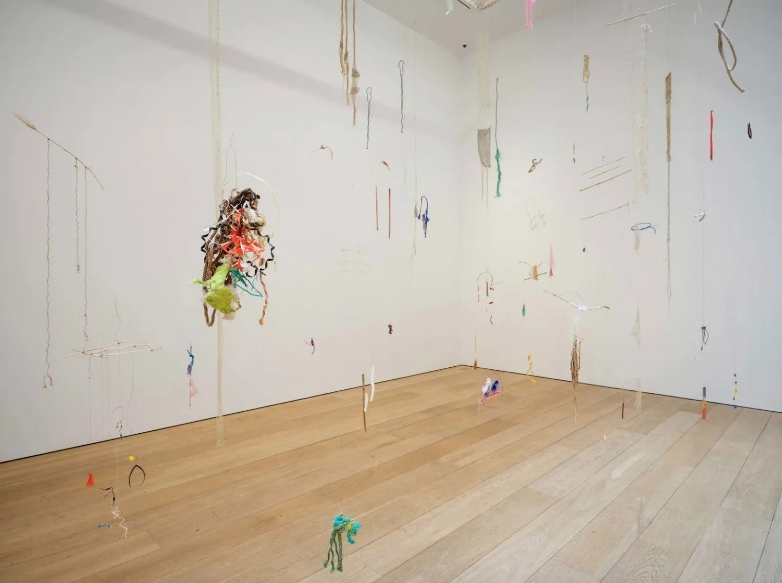 Installation view of a gallery, shot facing the intersection of two white walls on a light wood floor. Various colorful bits of objects varying in form hang around the room from invisible wire