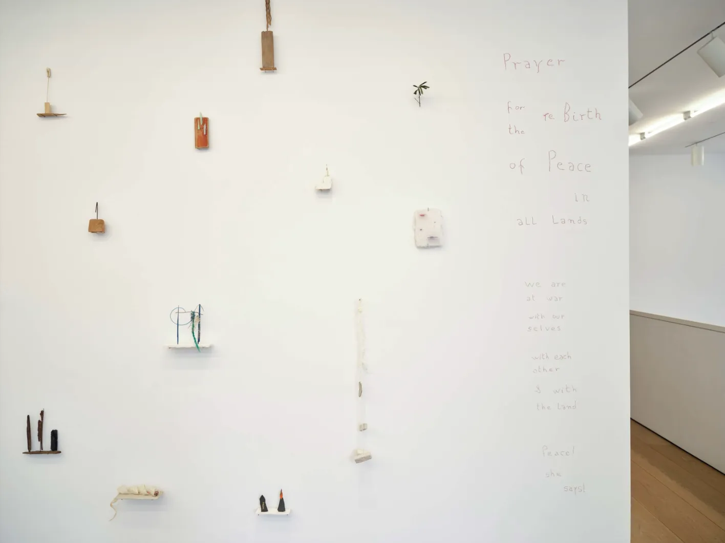 Installation view of gallery featuring a partial view of a free-standing white wall. On the left side of view, there are various object mounted in a seemingly random placement. To the right, near the edge of the wall, a column ofred handwritten text.