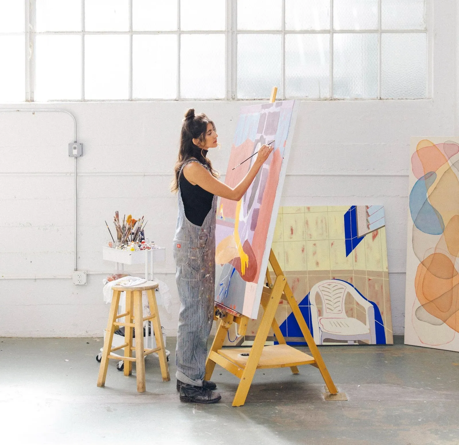 An artist wearing black top and jean overall painting in sun-lit studio with works in the background, indian artist Anoushka Mirchandani interview with shreya ajmani, impulse magazine.