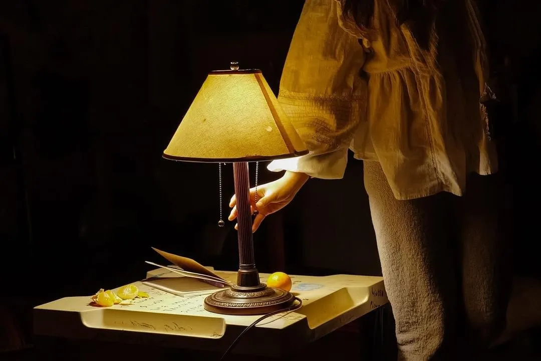 Against a dark, black background, a single table lamp illuminates a figure clad in white whose hand reaches for the lamp. On the small table, there appears to be a book, a peeled orange, and an orange ball. The desk has writing on its surface.
