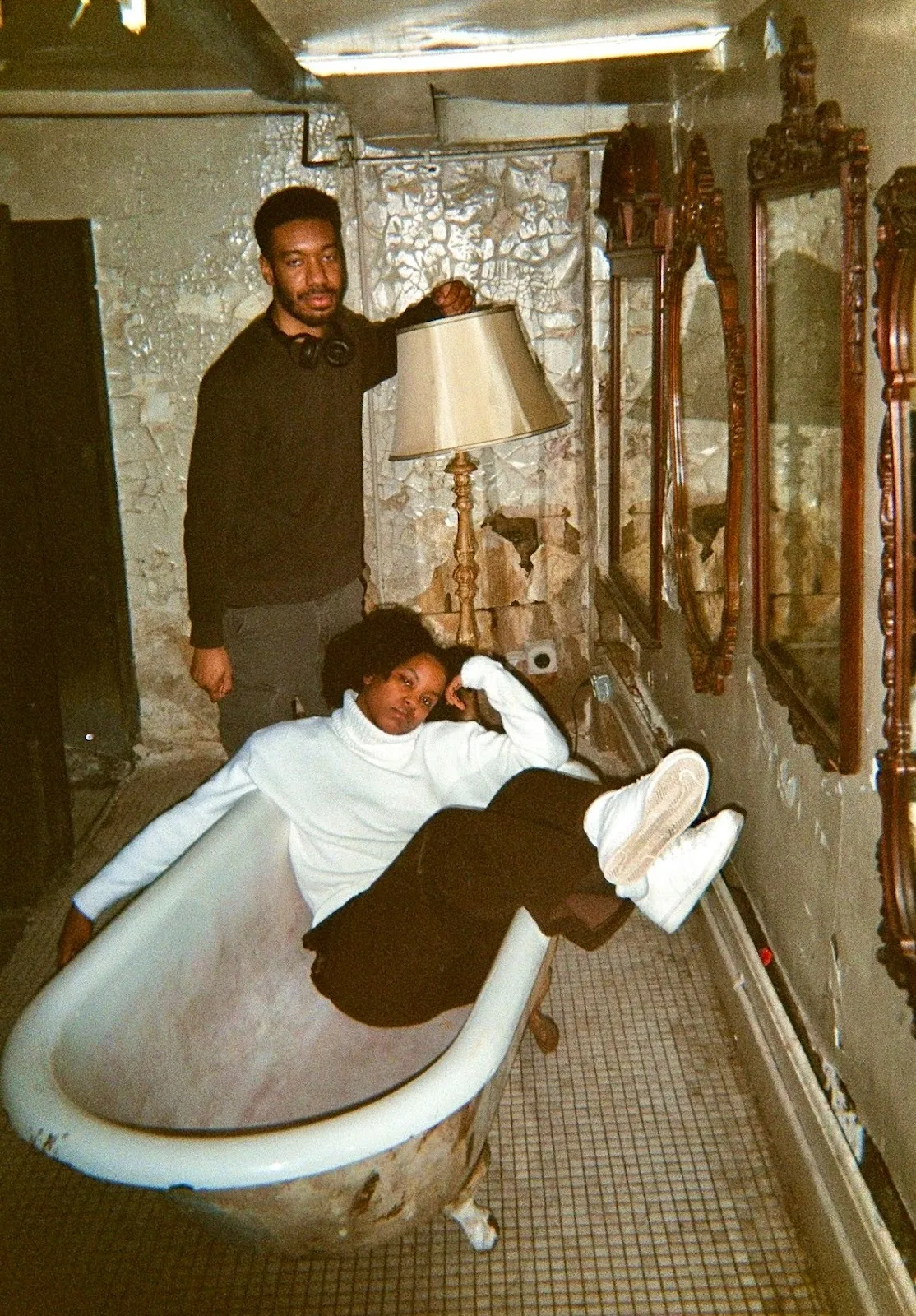 Two party goers in a Bushwick church basement bathroom, woman in white shirt lying inside the bathtub while a man stands behind her, next to rugged wall with mirrors, stamina, project iii nick brito collaboration techno night.